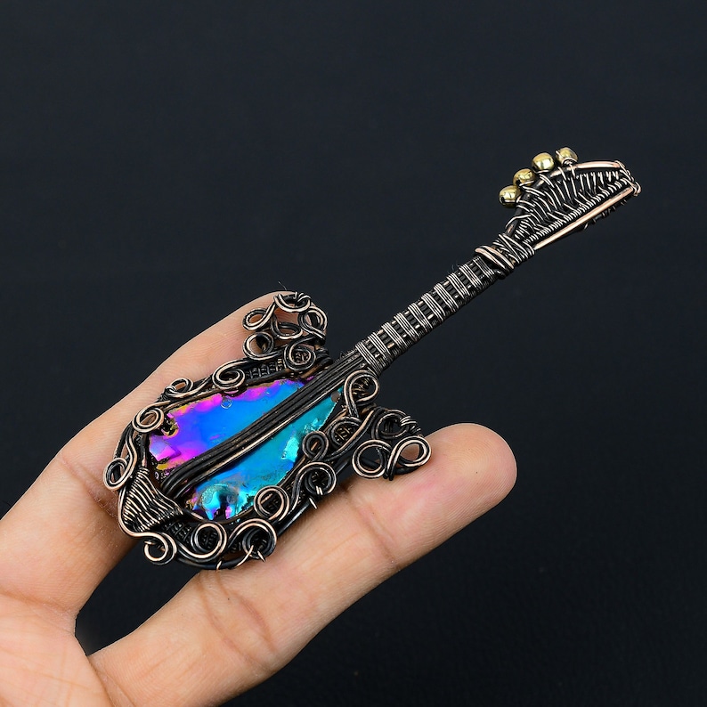 Puede incluir: Un colgante de guitarra envuelto en alambre con una piedra preciosa iridiscente en el cuerpo. La guitarra est&aacute; hecha de alambre de cobre y tiene un dise&ntilde;o detallado. El colgante es una pieza de joyer&iacute;a &uacute;nica y elegante.