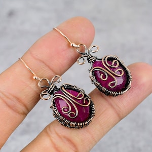 May include: A pair of handcrafted earrings featuring faceted, oval-shaped, deep magenta gemstones. The gemstones are encased in a copper wire frame with decorative swirls and a copper ear wire.