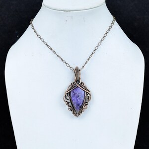 Peut inclure: Un collier pendentif cuivr&eacute; avec une pierre violette. La pierre est sertie dans un cadre orn&eacute;, enroul&eacute; de fil de fer. Le pendentif est suspendu &agrave; une cha&icirc;ne d&eacute;licate, cr&eacute;ant un bijou saisissant.