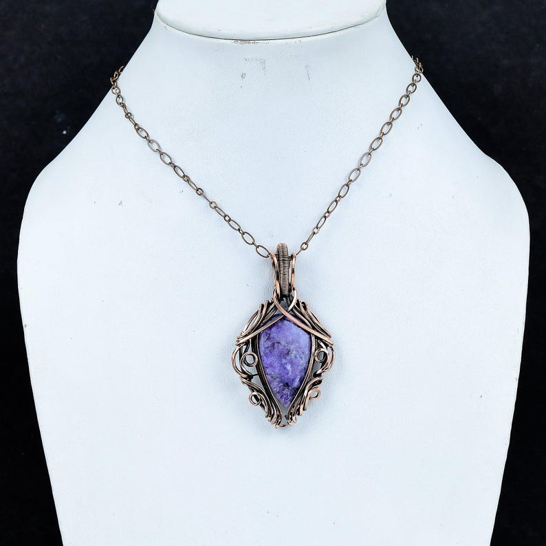 Peut inclure: Un collier pendentif cuivr&eacute; avec une pierre violette. La pierre est sertie dans un cadre orn&eacute;, enroul&eacute; de fil de fer. Le pendentif est suspendu &agrave; une cha&icirc;ne d&eacute;licate, cr&eacute;ant un bijou saisissant.