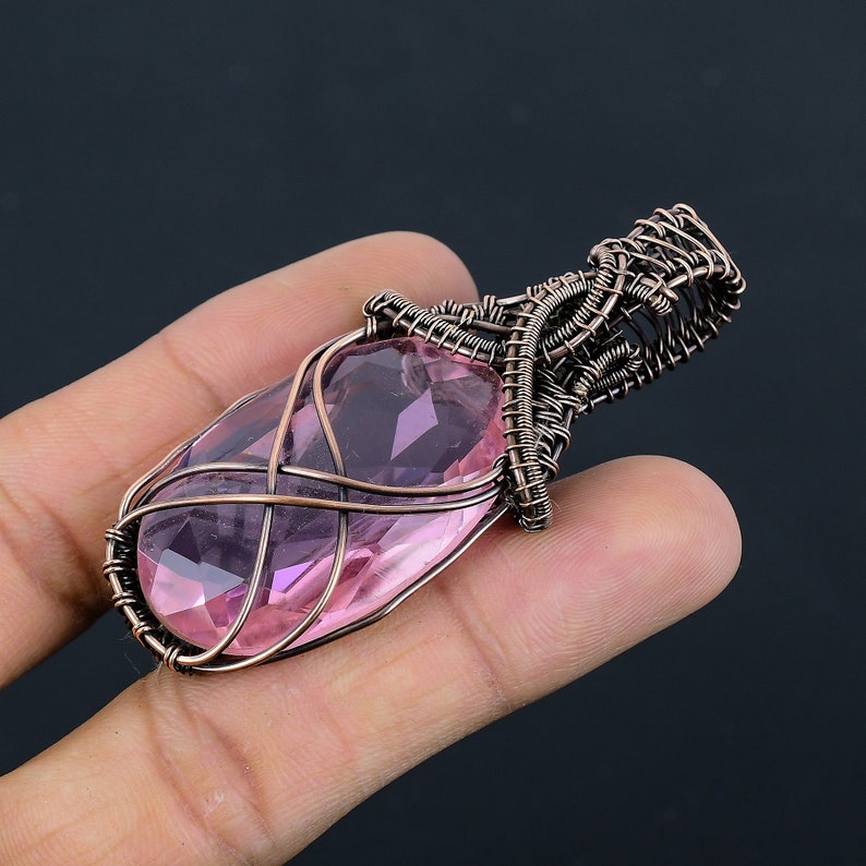 May include: A pink gemstone pendant wrapped in copper wire. The pendant is oval shaped and has a faceted design. The wire is wrapped around the stone in a decorative pattern.