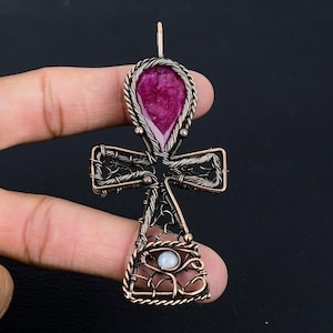 May include: A copper-toned pendant in the shape of an ankh cross. The top features a large, teardrop-shaped, deep pink gemstone. The cross arms and base are intricately wired, with a small, round, white gemstone at the base. The pendant is suspended from a small loop.