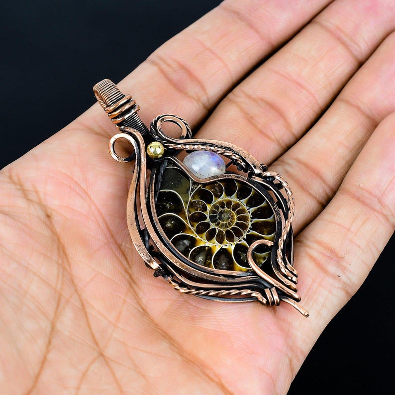 Puede incluir: Colgante artesanal con un f&oacute;sil de amonita encapsulado en alambre de cobre. El colgante tiene un dise&ntilde;o en espiral en tonos marrones y amarillos, con una peque&ntilde;a piedra lunar iridiscente y una cuenta dorada.