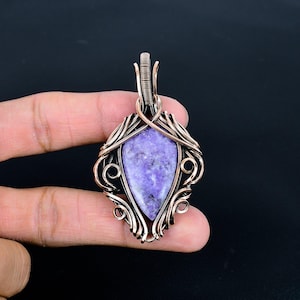 Peut inclure: Pendentif artisanal avec une pierre pr&eacute;cieuse violette en forme de goutte d'eau, sertie dans une monture en fil de cuivre. Le filigrane pr&eacute;sente des volutes et des courbes complexes, avec une b&eacute;li&egrave;re en haut pour une cha&icirc;ne. Le pendentif est sur fond sombre.