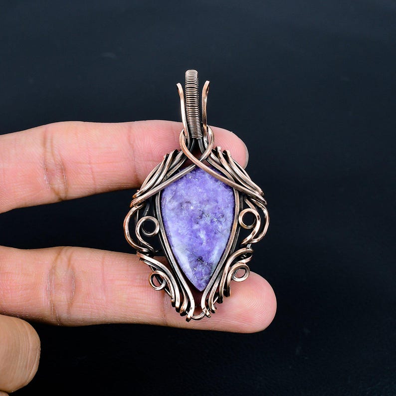 Peut inclure: Pendentif artisanal avec une pierre pr&eacute;cieuse violette en forme de goutte d'eau, sertie dans une monture en fil de cuivre. Le filigrane pr&eacute;sente des volutes et des courbes complexes, avec une b&eacute;li&egrave;re en haut pour une cha&icirc;ne. Le pendentif est sur fond sombre.