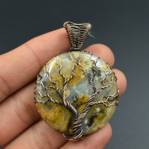 Puede incluir: Un colgante de &aacute;rbol de la vida envuelto en alambre de cobre con una piedra de &aacute;gata redonda de color amarillo, marr&oacute;n y blanco.
