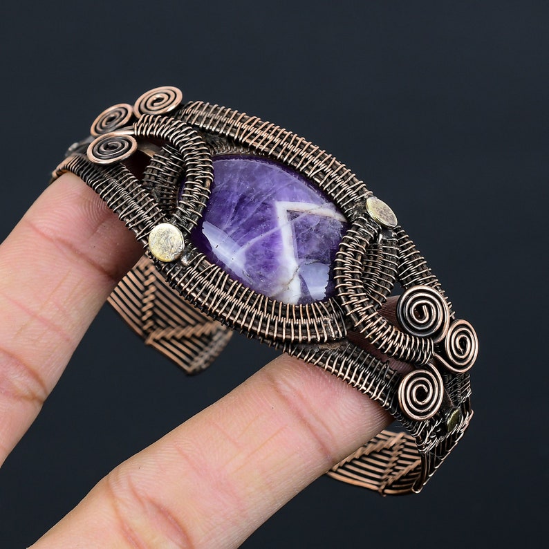 May include: A handcrafted copper wire cuff bracelet featuring a large, oval-shaped amethyst stone. The amethyst is a deep purple with white inclusions. The bracelet has intricate wirework and decorative spiral accents. The cuff is held in a hand.