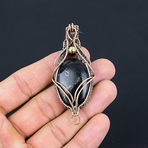 May include: A black gemstone pendant with a copper wire wrap design. The pendant has a gold bead at the top and a loop at the bottom for attaching a chain.