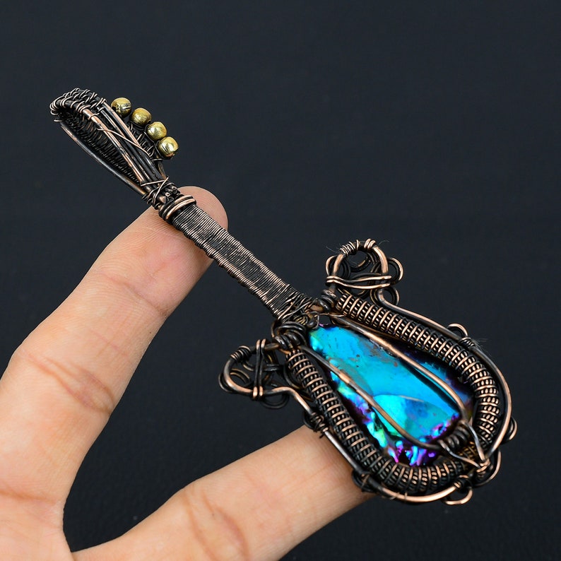 Puede incluir: Un colgante de guitarra envuelto en alambre con una piedra preciosa azul iridiscente. El colgante est&aacute; hecho de alambre de cobre y tiene un dise&ntilde;o detallado.