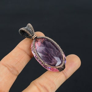 Puede incluir: Un colgante de piedra preciosa rosa con talla ovalada, encerrado en un marco de alambre de cobre. El alambrismo forma un borde y una argolla decorativos. El colgante se sostiene en una mano sobre un fondo oscuro.