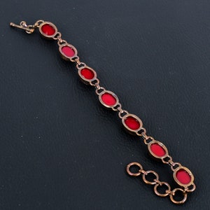 K&ouml;nnte beinhalten: Ein Kupferarmband mit roten ovalen Steinen. Das Armband hat einen Karabinerverschluss und eine Kettenverl&auml;ngerung.