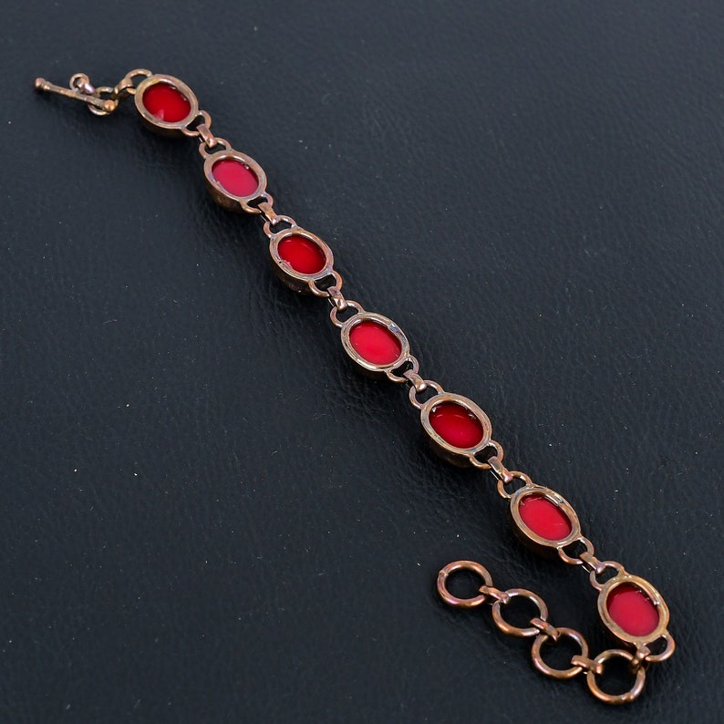 K&ouml;nnte beinhalten: Ein Kupferarmband mit roten ovalen Steinen. Das Armband hat einen Karabinerverschluss und eine Kettenverl&auml;ngerung.