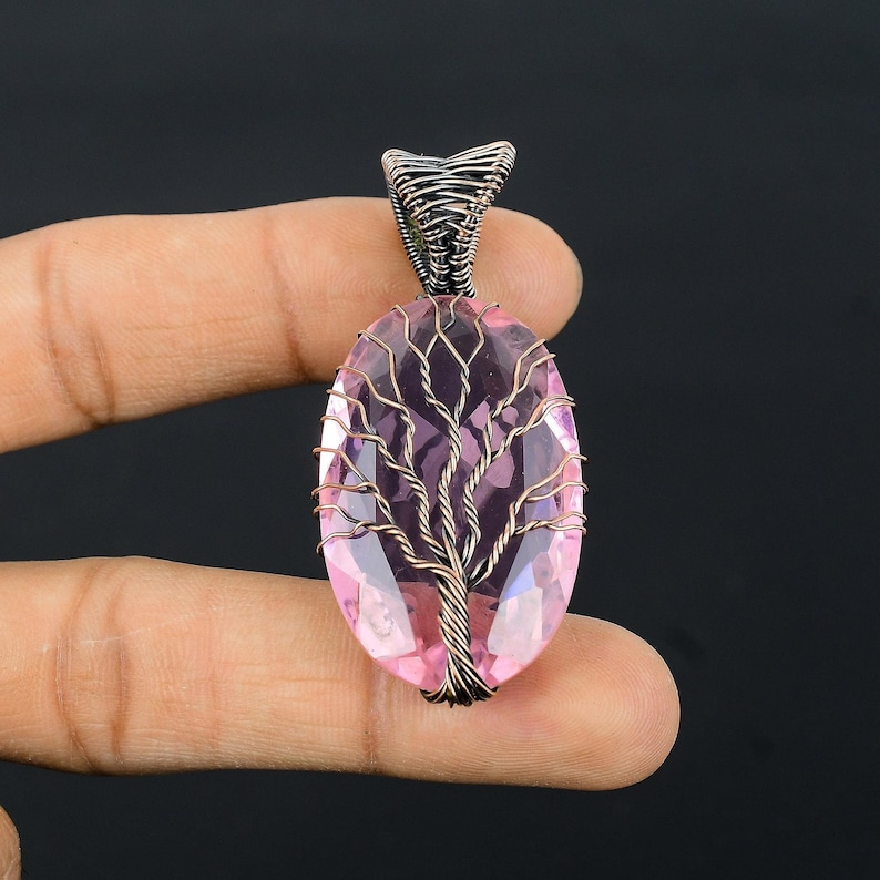 Puede incluir: Colgante artesanal con una piedra preciosa rosa de forma ovalada, con un dise&ntilde;o de &aacute;rbol envuelto en alambre. El colgante tiene una argolla plateada y lo sostiene una mano sobre un fondo oscuro. La piedra mide aproximadamente 3,8 cm de largo.