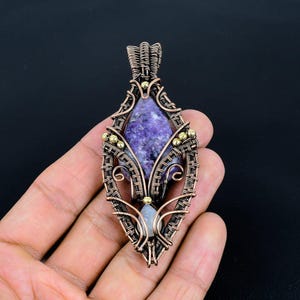 May include: A handcrafted pendant featuring a purple gemstone set within a copper wire frame. The wire is intricately woven, with small gold-colored beads adding detail. The pendant has a teardrop shape.