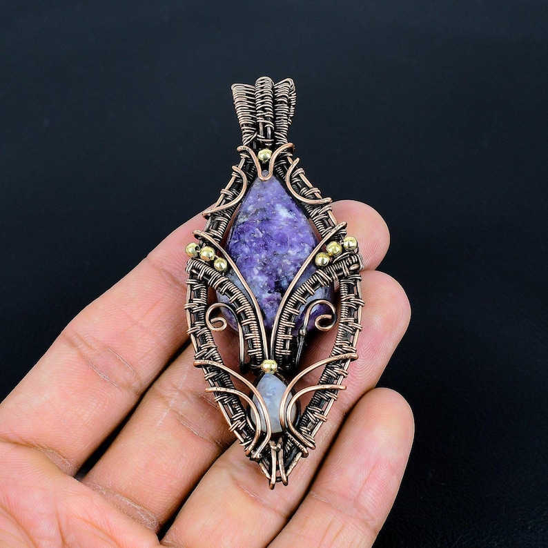May include: A handcrafted pendant featuring a purple gemstone set within a copper wire frame. The wire is intricately woven, with small gold-colored beads adding detail. The pendant has a teardrop shape.