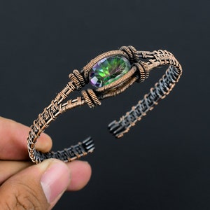 May include: A copper wire wrapped bracelet with a green and iridescent gemstone set in the center. The bracelet is made with a woven wire design.