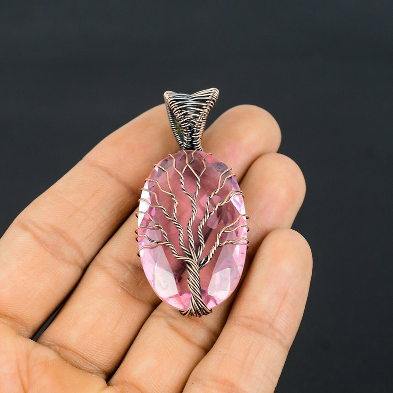 Puede incluir: Un colgante de piedra preciosa rosa con un dise&ntilde;o de &aacute;rbol envuelto en alambre. La piedra ovalada es de color rosa claro, y el &aacute;rbol est&aacute; hecho de alambre color cobre. La fianza est&aacute; hecha de alambre m&aacute;s oscuro y apretado. El colgante est&aacute; sobre un fondo oscuro.