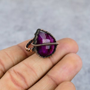 May include: A handcrafted ring featuring a pear-shaped, faceted, deep magenta gemstone. The stone is encased in a dark, textured metal setting, with a copper-colored twisted band. The ring is displayed on a light gray background.