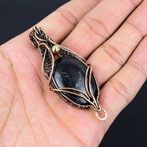May include: A black gemstone pendant with copper wire wrapping and a gold bead accent. The pendant is oval shaped and has a wire loop for attaching to a chain.