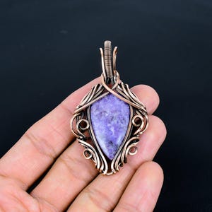 Peut inclure: Un pendentif en cuivre fait &agrave; la main avec une pierre en forme de goutte d'eau de couleur lavande. La pierre est sertie dans un fil de cuivre complexe avec des volutes d&eacute;coratives et une b&eacute;li&egrave;re pour une cha&icirc;ne. Le pendentif est tenu dans une main sur fond sombre.
