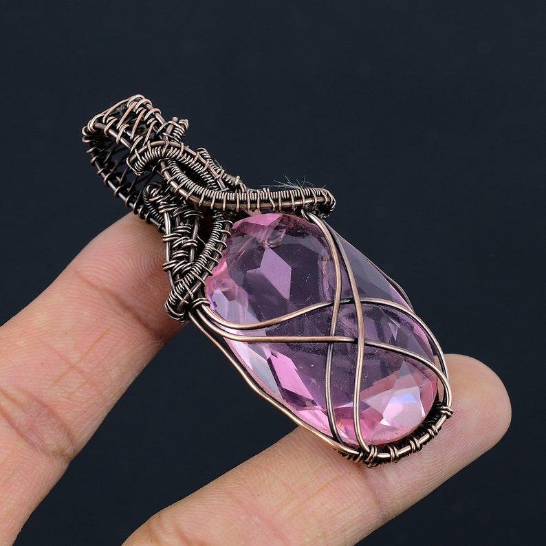 May include: A pink crystal pendant wrapped in copper wire. The pendant is oval shaped and has a faceted design. The wire is wrapped around the crystal in a decorative pattern.