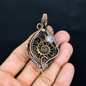 Puede incluir: Colgante envuelto en alambre de cobre con un caparaz&oacute;n de amonita fosilizada con un patr&oacute;n en espiral. El colgante incluye una peque&ntilde;a piedra preciosa azul claro y una cuenta dorada. La amonita est&aacute; encerrada en un marco de alambre de cobre.