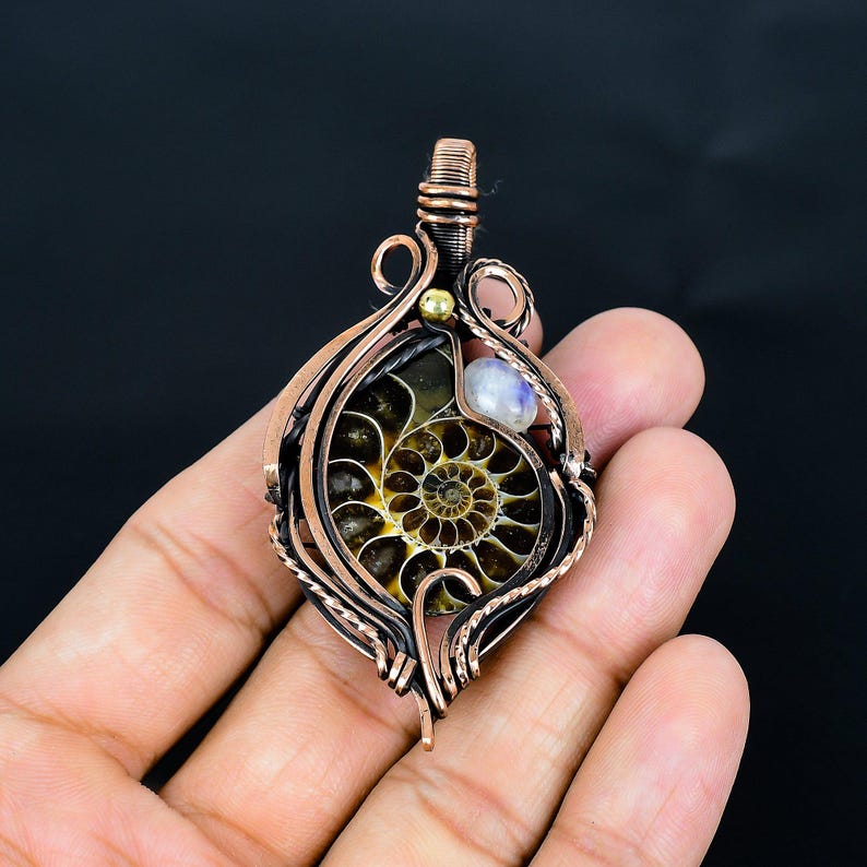 Puede incluir: Colgante envuelto en alambre de cobre con un caparaz&oacute;n de amonita fosilizada con un patr&oacute;n en espiral. El colgante incluye una peque&ntilde;a piedra preciosa azul claro y una cuenta dorada. La amonita est&aacute; encerrada en un marco de alambre de cobre.