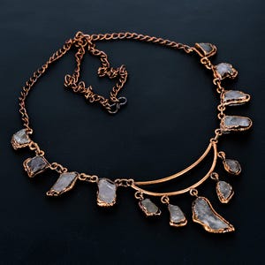 May include: A copper-colored necklace with a crescent moon design. The necklace features irregularly shaped, light-colored stones with gold-colored edges. The stones are connected by a delicate chain, creating a unique and elegant accessory.