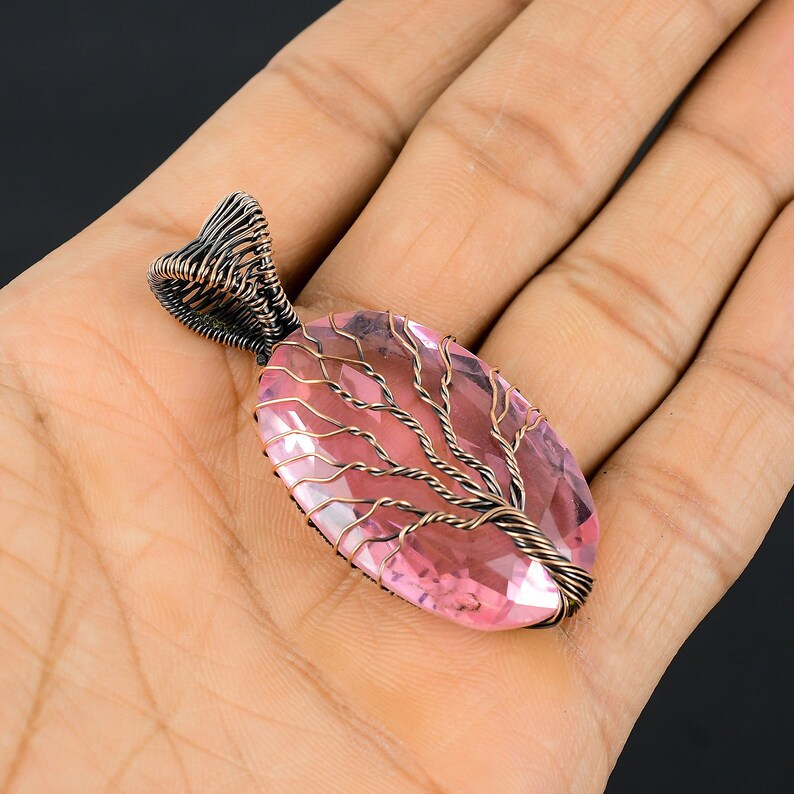 Puede incluir: Un colgante artesanal con una gran gema rosa ovalada. La piedra est&aacute; engastada en un dise&ntilde;o de alambre color cobre que se asemeja a un &aacute;rbol. La argolla tambi&eacute;n est&aacute; envuelta en alambre. El colgante se sostiene en una mano sobre un fondo oscuro.