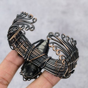 May include: A black and copper wire wrapped bracelet with a large, dark grey stone in the center. The bracelet has a decorative design of swirls and loops.