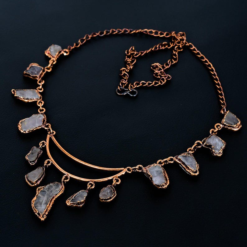 May include: A rose gold necklace featuring irregularly shaped, light gray gemstones. The stones are set in a gold-colored metal and are connected by a delicate chain. The necklace has a crescent-shaped design with stones hanging from the bottom.