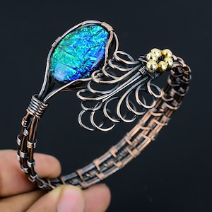 Puede incluir: Un brazalete de alambre de cobre con un cabujón de vidrio dicroico azul y verde y cuentas doradas. El brazalete está hecho con un diseño de alambre tejido.