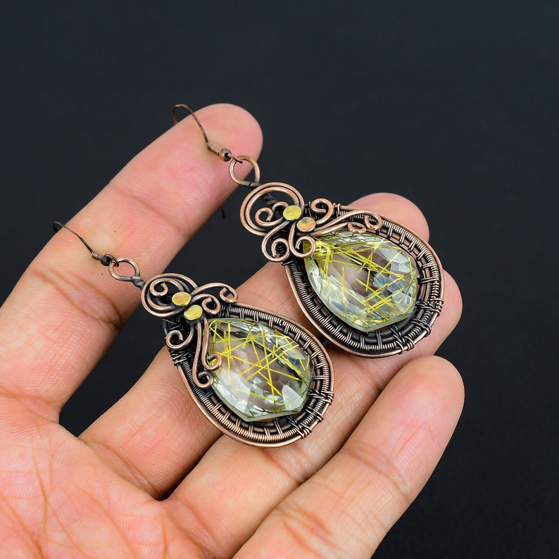 May include: A pair of teardrop-shaped earrings with a green and gold crystal stone wrapped in copper wire. The earrings have a delicate, intricate design with a floral motif.