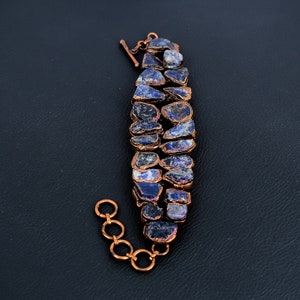 K&ouml;nnte beinhalten: Ein Kupferarmband mit einer Reihe von blauen und violetten Edelsteinen. Das Armband hat einen Karabinerverschluss.