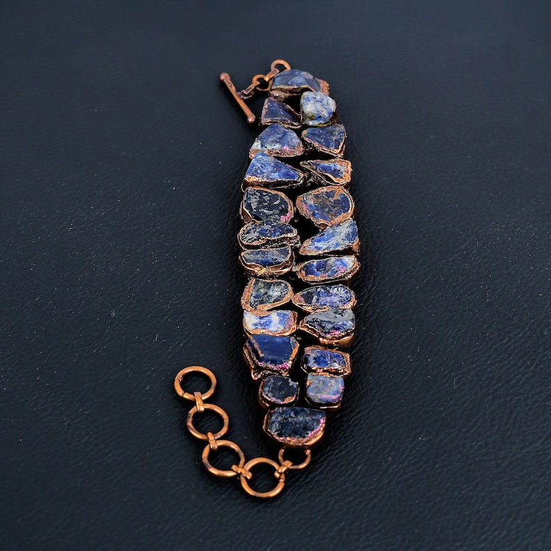 K&ouml;nnte beinhalten: Ein Kupferarmband mit einer Reihe von blauen und violetten Edelsteinen. Das Armband hat einen Karabinerverschluss.