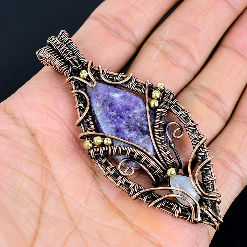 May include: A handcrafted pendant featuring a large, oval-shaped purple stone, intricately wrapped in copper wire. Small gold-colored beads accent the design. A smaller, white stone is set at the bottom. The pendant is held in a hand.