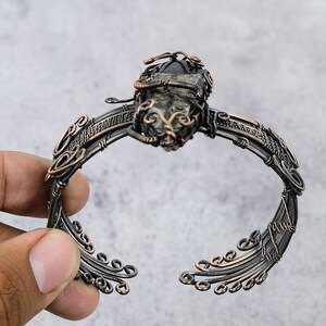 May include: A copper wire wrapped bracelet with a dark gray gemstone in the center. The bracelet has a delicate, intricate design with swirling patterns.