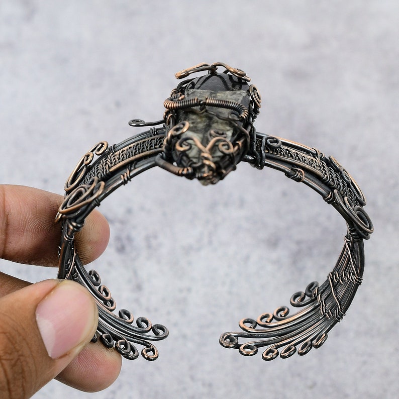 May include: A copper wire wrapped bracelet with a dark gray gemstone in the center. The bracelet has a delicate, intricate design with swirling patterns.