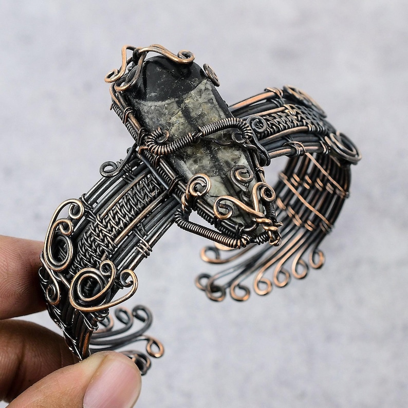 May include: A copper wire wrapped bracelet with a black and grey stone set in the center. The bracelet is made with intricate wirework and has a delicate, feminine design.