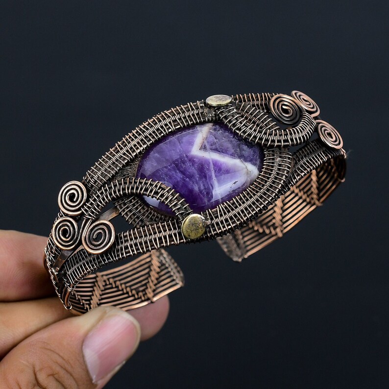 May include: A handcrafted copper wire cuff bracelet featuring a large, oval amethyst gemstone. The bracelet has intricate wirework with spiral accents and small brass-colored rivets. The amethyst is a deep purple color with white veining.