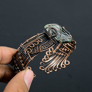 May include: A copper wire wrapped bracelet with a green and black stone set in the center. The bracelet is made with intricate wirework and has a delicate, flowing design.