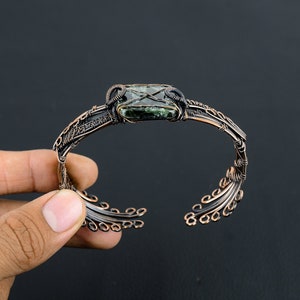 May include: A copper wire wrapped bracelet with a green and black stone set in the center. The bracelet has a delicate, intricate design.