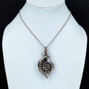 Puede incluir: Collar con colgante de alambre color cobre con un amonita fosilizada. El colgante tiene forma de l&aacute;grima con un dise&ntilde;o en espiral, una peque&ntilde;a perla y una piedra lunar. El collar se exhibe en un maniqu&iacute; blanco.