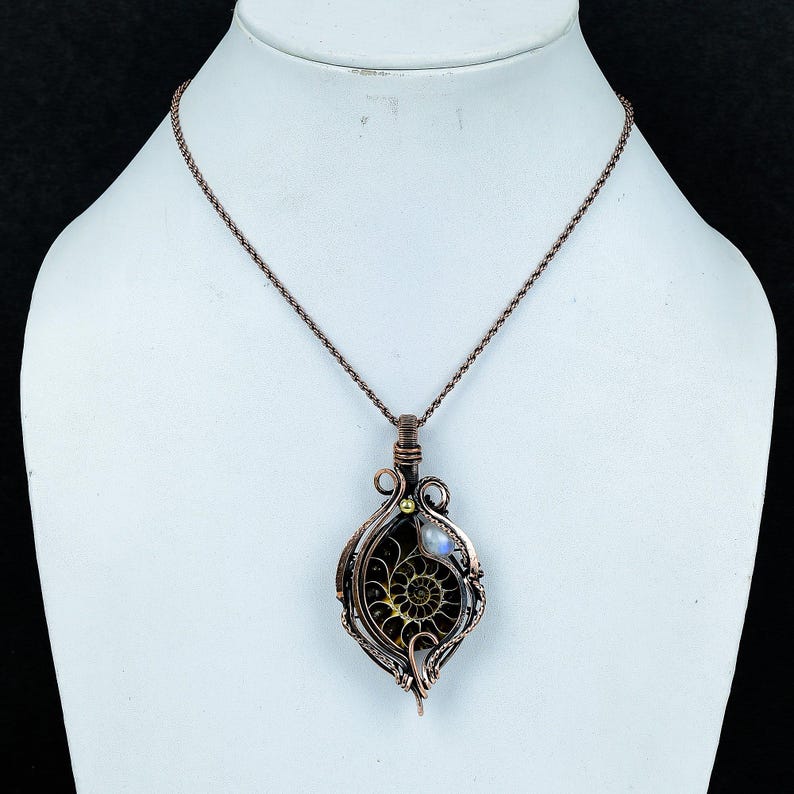 Puede incluir: Collar con colgante de alambre color cobre con un amonita fosilizada. El colgante tiene forma de l&aacute;grima con un dise&ntilde;o en espiral, una peque&ntilde;a perla y una piedra lunar. El collar se exhibe en un maniqu&iacute; blanco.