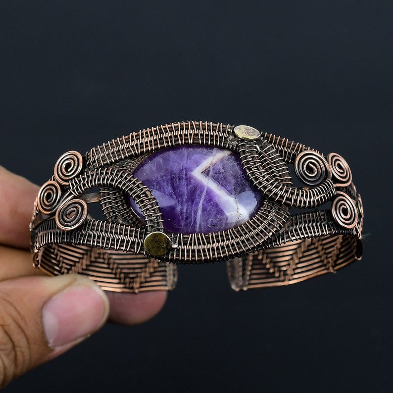 May include: A handcrafted copper cuff bracelet featuring a large, oval amethyst stone. The stone is a deep purple with white veining. The bracelet has intricate wirework and decorative spiral accents.
