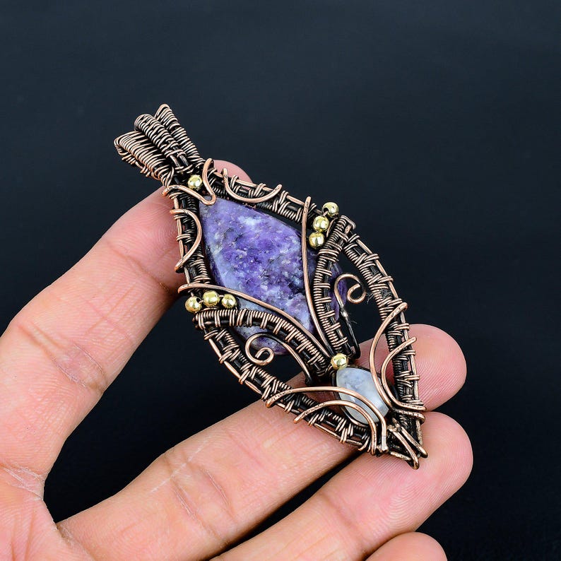 May include: A handcrafted pendant featuring a central, oval-shaped purple stone, intricately wrapped in copper wire. Small gold-colored beads accent the design. The pendant has a teardrop shape and a small, light-colored stone at the bottom.