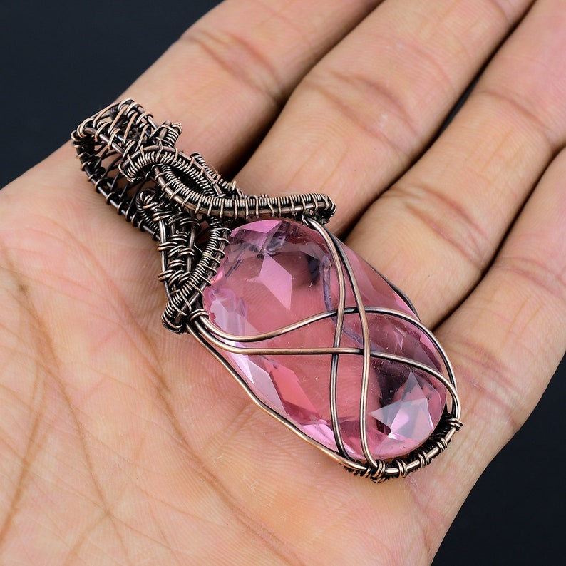 May include: A pink crystal pendant wrapped in copper wire. The pendant is oval shaped and has a faceted design. The wire is woven into a delicate pattern around the crystal.