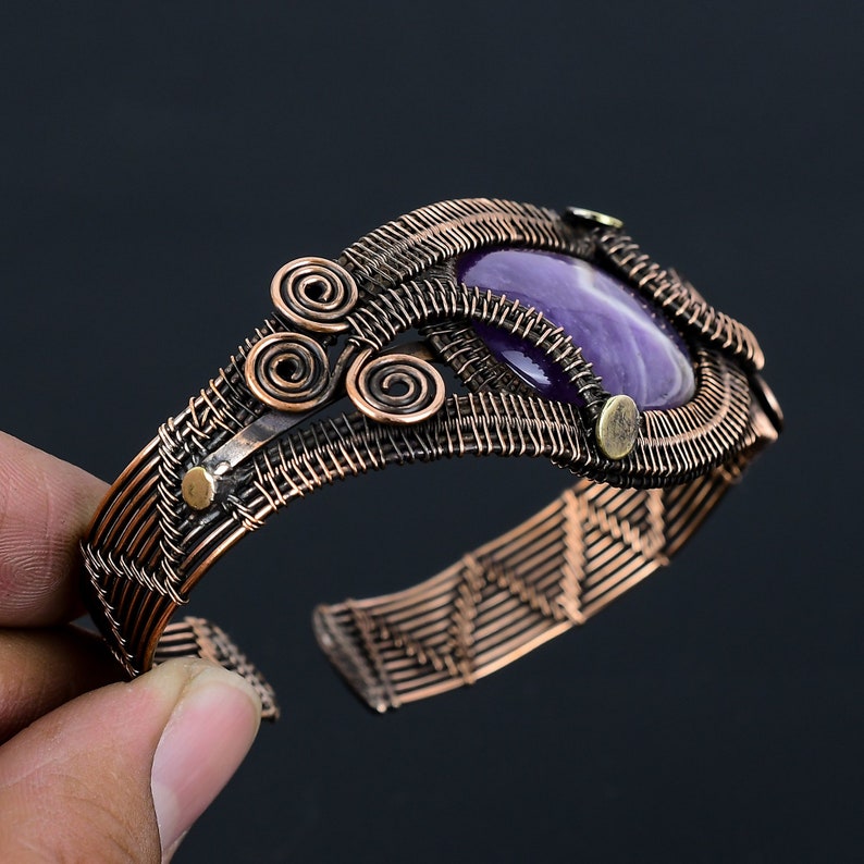 May include: A handcrafted copper cuff bracelet featuring a large, oval-shaped purple amethyst stone. The bracelet has intricate wirework with spiral and woven patterns, and small brass accents. The open design allows for easy wear.