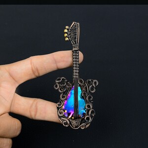 Puede incluir: Un colgante de guitarra de alambre de cobre con una piedra iridiscente azul y p&uacute;rpura en el centro. El colgante est&aacute; sostenido por un dedo sobre un fondo negro.