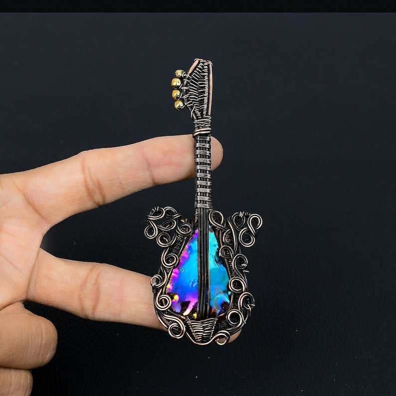 Puede incluir: Un colgante de guitarra de alambre de cobre con una piedra iridiscente azul y p&uacute;rpura en el centro. El colgante est&aacute; sostenido por un dedo sobre un fondo negro.